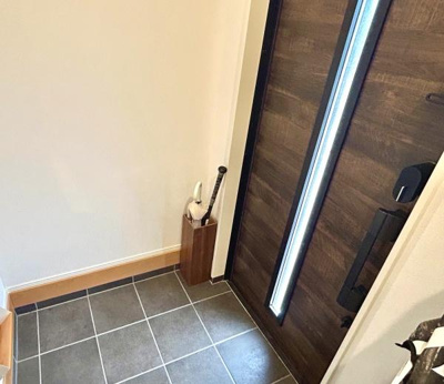 玄関扉は木目調のデザインで落ち着いた雰囲気を感じられます。タイル張りの床が清潔感を演出しています。明るさと開放感のある玄関スペースです。シンプルな内装が特徴となっています。