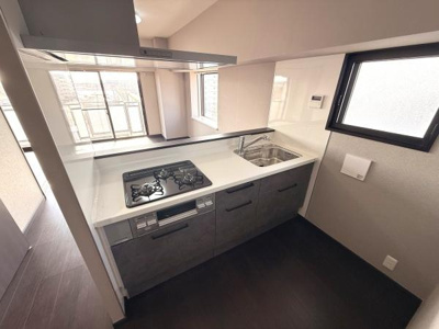 窓が複数あり明るい空間が広がるキッチンです。シンプルなデザインで使いやすさが感じられます。三口コンロと大きなシンクが配置されています。収納が充実しているのも特徴です。全体的にすっきりとした印象です。