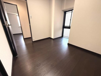 ダークカラーのフローリングが落ち着いた印象を与える洋室です。窓からの自然光が室内を明るく照らします。建具や扉もシンプルなデザインで統一感のある空間です。隣室との繋がりも感じられる間取りです。