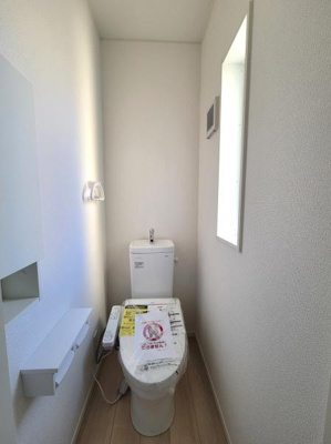 窓から自然光が入り明るい印象のトイレです。ホワイトを基調とした内装で清潔感があります。棚や収納スペースも配置され使いやすさが考慮されています。空間が縦長で圧迫感の少ないデザインです。