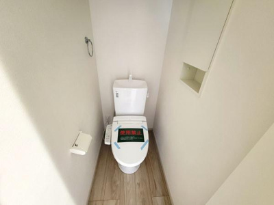 シンプルなデザインのトイレです。明るい色合いの内装で清潔感があります。壁面に収納スペースがあり使い勝手の良さを感じられます。床は木目調の仕上げです。アクセントになるタオル掛けも設置されています。