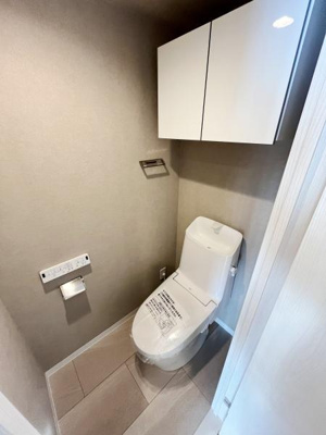 シンプルなデザインのトイレです。手の届きやすい位置に収納棚が設置されています。壁付けのペーパーホルダーが使い勝手を向上させています。温水洗浄便座が備わっています。清潔感のある空間です。