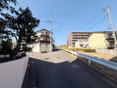 道路が広く車の通行がしやすい印象です。周囲には複数の住宅やマンションが立ち並んでいます。ガードレールが設置されており安全面にも配慮されています。