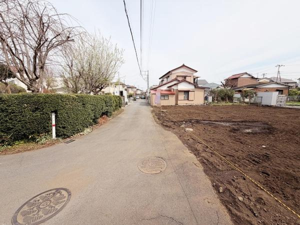 現地写真で前面道路の様子がよく分かります。周囲には家や植栽があり落ち着いた環境となっています。道路幅も確認しやすい写真です。前面道路の舗装状態も一目でわかります。