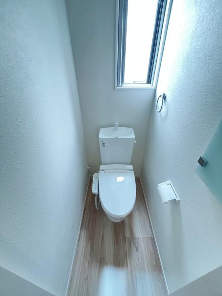 明るい光が差し込むトイレ空間です。清潔感のある白基調の内装が特徴的です。窓があり換気もしやすい設計になっています。床は木目調で落ち着いた雰囲気です。