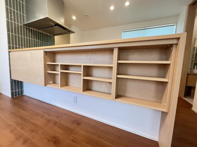 リビングとキッチンの間に設置された収納棚が空間を有効に活用しています。木目調の仕上がりが室内と調和しています。清潔感のある床材が印象的です。天井のダウンライトが明るさを確保しています。