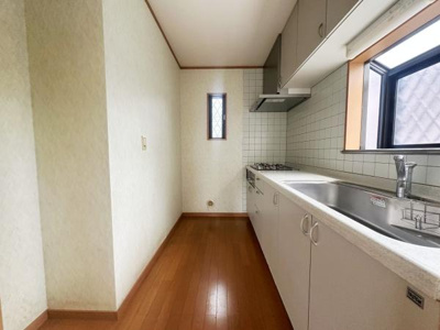 窓から自然光が差し込む明るいキッチンです。シンプルなタイルの壁面と広い作業スペースが特徴です。床は木目調で温かみを感じられます。換気窓もあり快適にお使いいただけます。