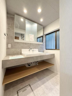 大きな鏡と広々としたカウンターが特徴の洗面台です。照明が明るく洗面スペースがゆったりしています。窓から自然光も差し込み清潔感があります。下部の棚は収納に便利です。無駄のないシンプルなデザインです。