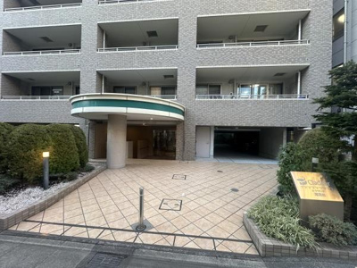 タイル張りのアプローチが特徴的なエントランスです。エントランス横にサインが設置されています。屋根付きで雨の日も安心して出入りできます。緑に囲まれた印象のスペースです。